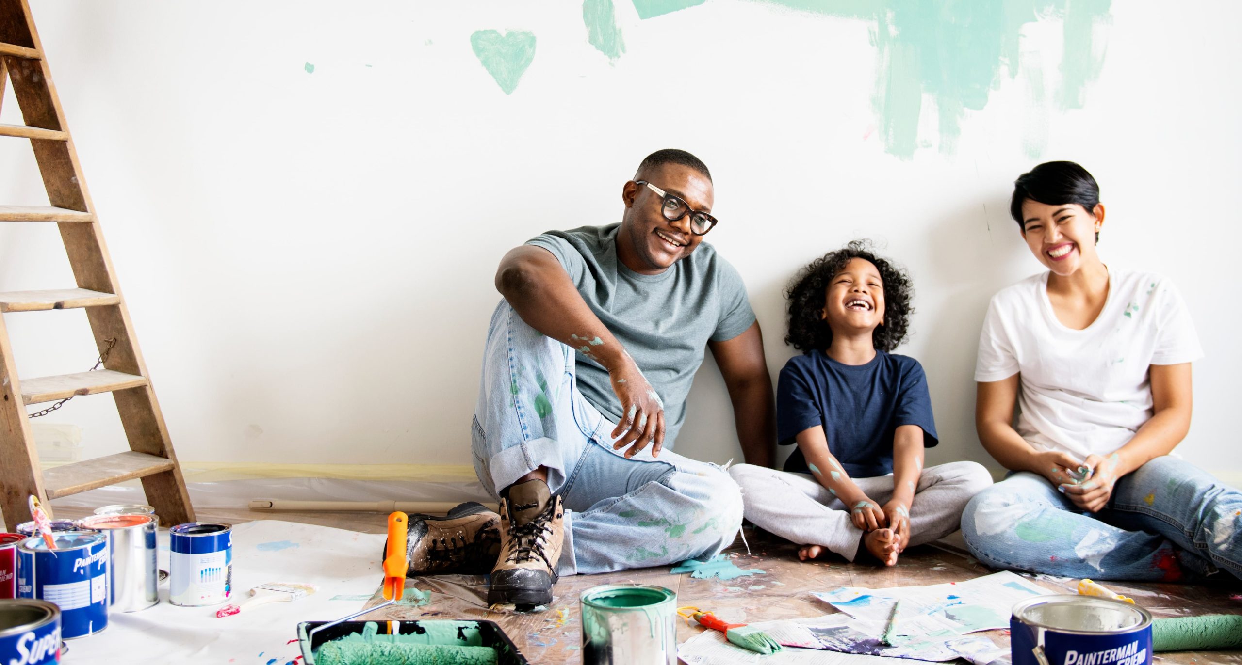 Couple with child sitting on floor laughing in room that they are painting